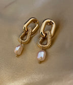 Close-up of sculptural gold link earrings featuring a twisted rope texture and hanging freshwater baroque pearl drops. The earrings are displayed on a champagne silk fabric to highlight the pearl luster and high-shine gold finish