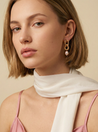 A female model wearing chunky gold interlocking link earrings with pearl drops. The vintage-inspired jewelry creates an elegant statement against a soft ivory background, styled with a pink silk camisole and white scarf