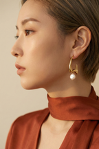 Close-up of a person wearing a gold hoop earring with a pearl, against a neutral background.