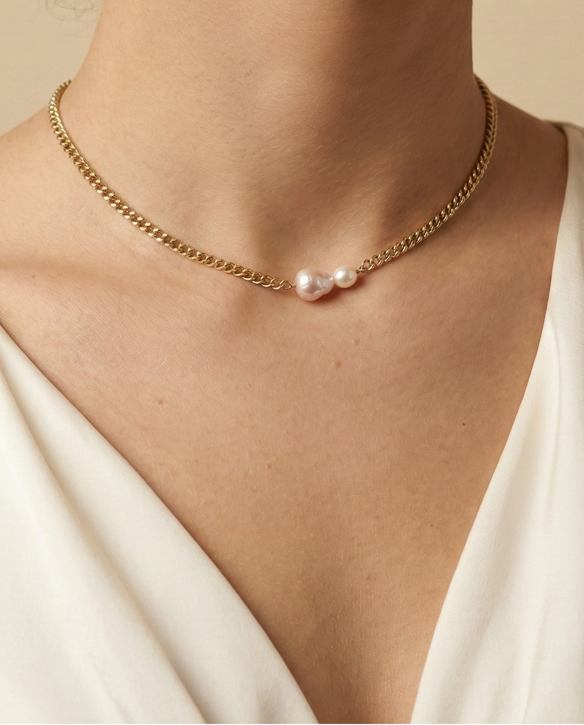 Close-up of a model wearing a dainty gold curb chain necklace featuring a pink baroque pearl and a small champagne pearl, styled with an ivory silk top