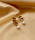 Close-up of gold chain link earrings featuring a lustrous oval white pearl drop. The earrings are shown on a model to demonstrate scale, featuring a high-polish gold finish and organic pearl shape