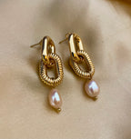 Close-up of sculptural gold link earrings featuring a twisted rope texture and hanging pink freshwater baroque pearl drops. The earrings are displayed on a champagne silk fabric to highlight the pearl luster and high-shine gold finish