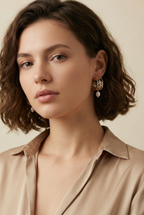 Close-up editorial profile of a model wearing detailed gold openwork hoop earrings featuring a leaf motif and a single dangling white oval pearl. The warm, soft lighting highlights the pearl's luster and the gold texture against the model's skin, set against a neutral background.