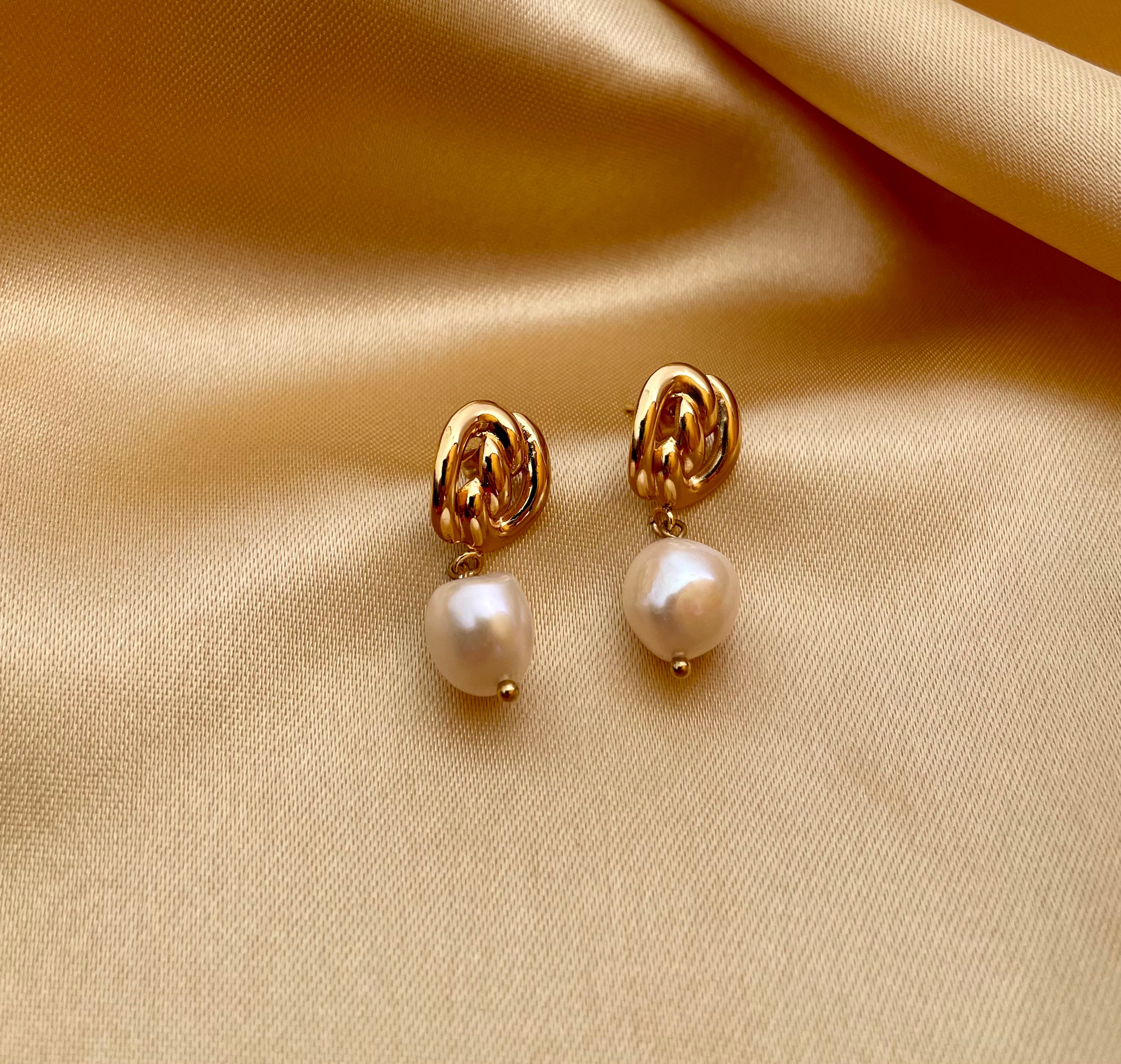 Close-up detail gold twist-knot stud earrings with a lustrous white pearl drop. The lighting highlights the texture of the gold and the glow of the pearl