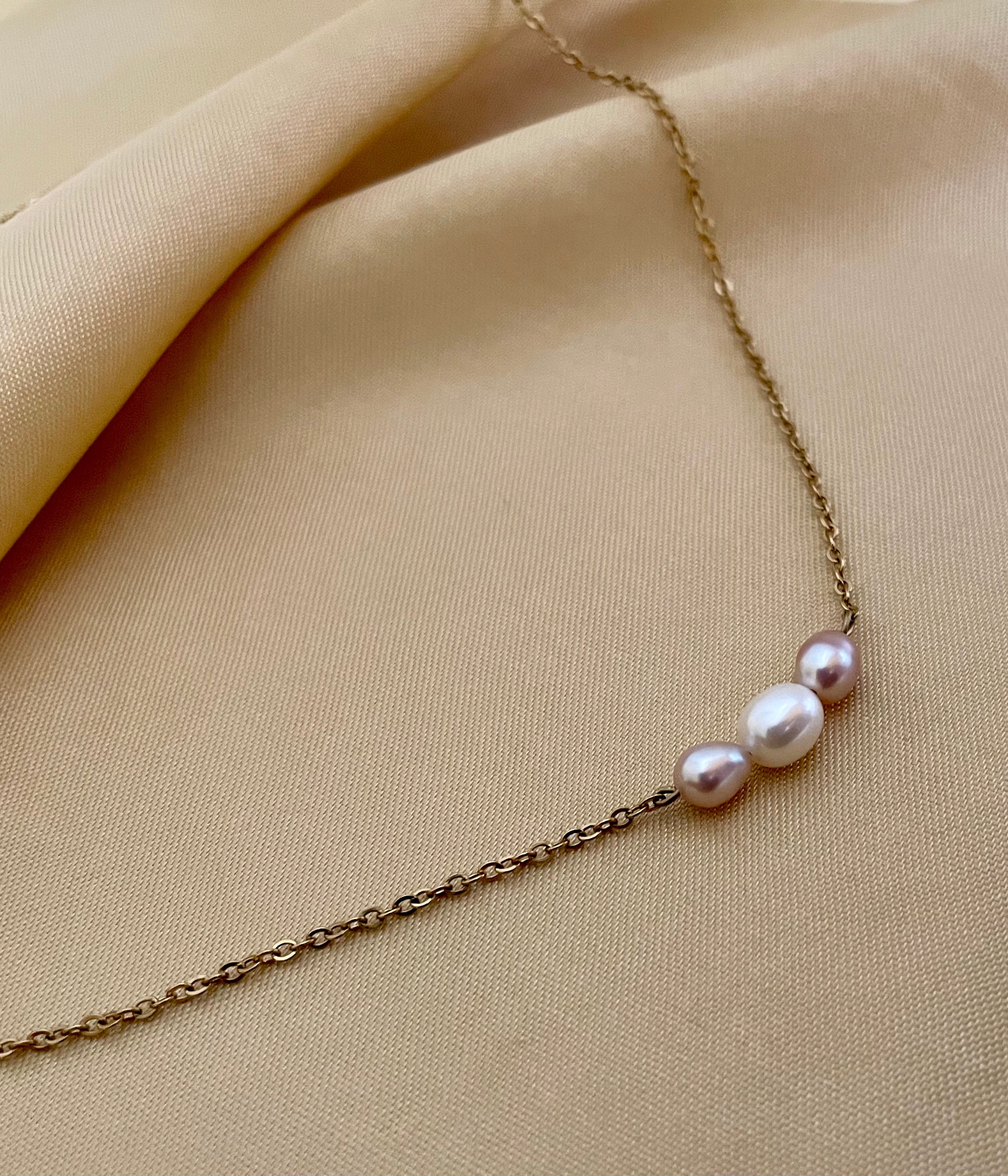 Close-up of a model wearing a dainty gold chain necklace featuring a trio of small pearls: a central white pearl flanked by two pink oval baroque pearls