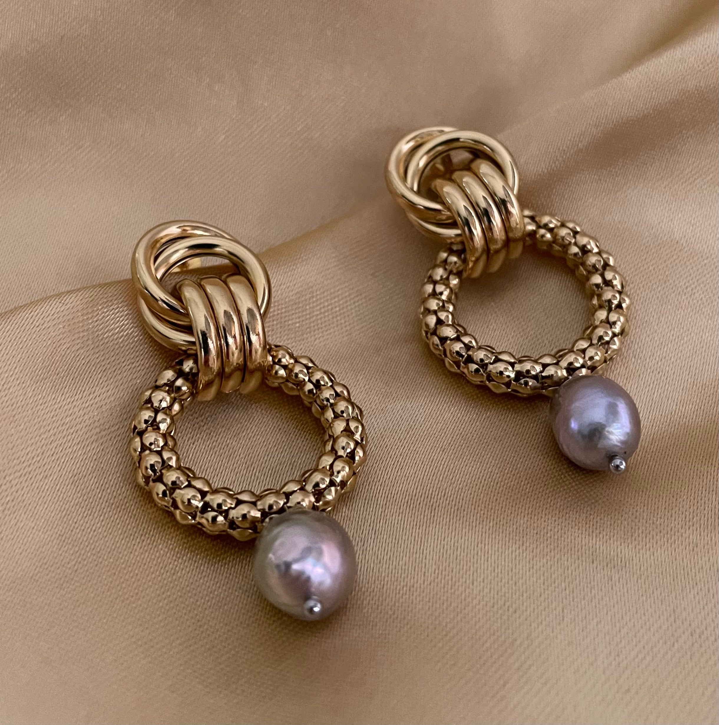 Close-up detail of textured gold interlocking hoop earrings featuring a lustrous metallic grey pearl drop. The design includes a beaded gold circle and polished gold links