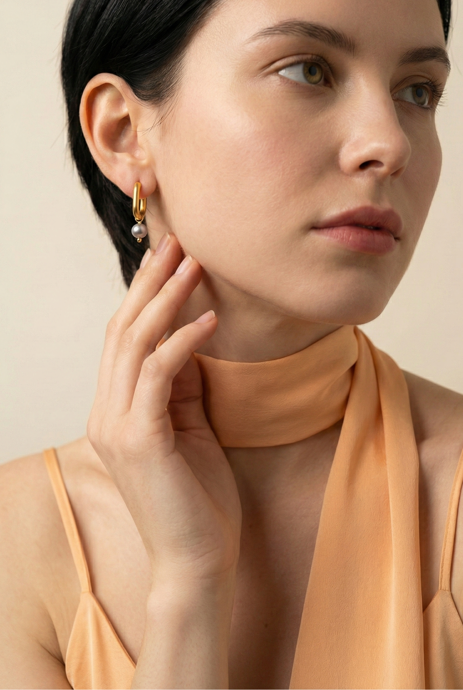 Editorial jewelry portrait showing a side profile of a model wearing luxury gold pearl earrings styled with an orange silk scarf.