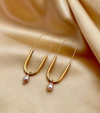 Close-up of a model wearing modern gold safety pin drop earrings featuring a lustrous pink oval pearl charm. Minimalist gold wire earrings with a U-shape design and natural pearl detail