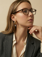Elegant side-profile of a blonde model wearing sophisticated gold and grey pearl drop earrings paired with an unbuttoned ivory blouse.