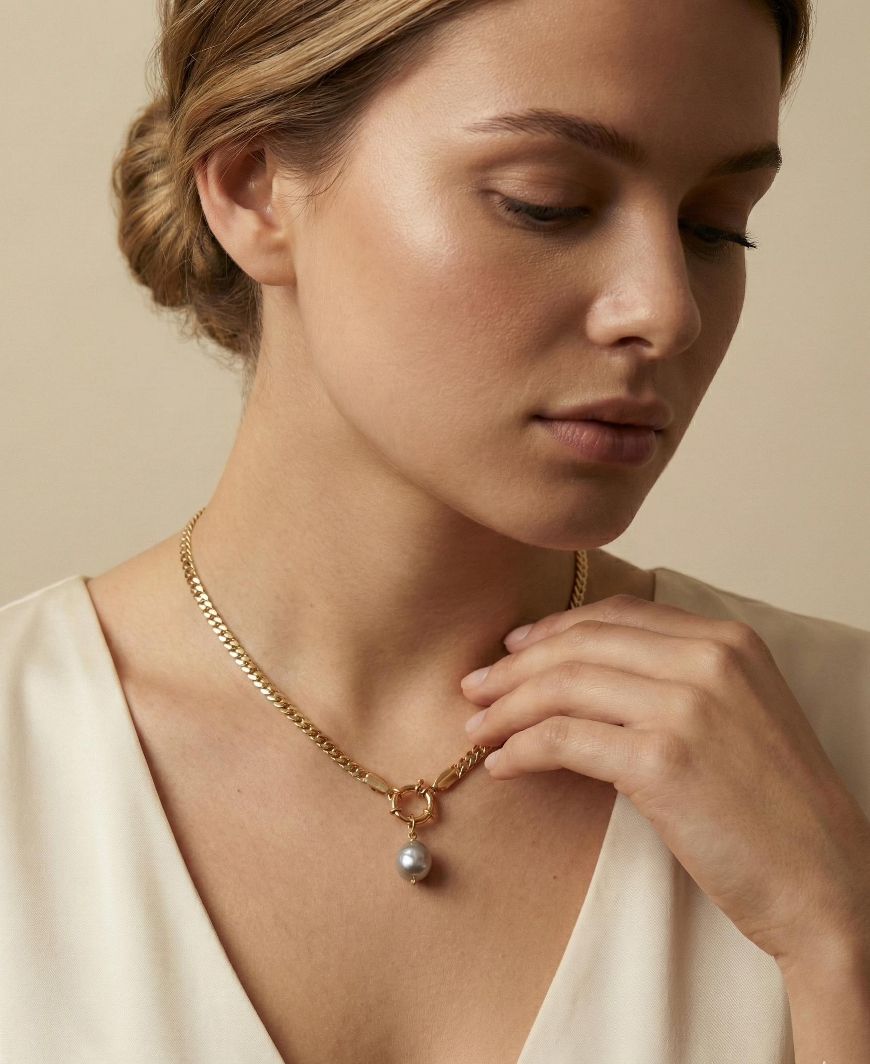 Close-up editorial shot of a woman wearing a minimalist gold chain with a grey baroque pearl drop, highlighting the necklace length and luster against warm skin tones.