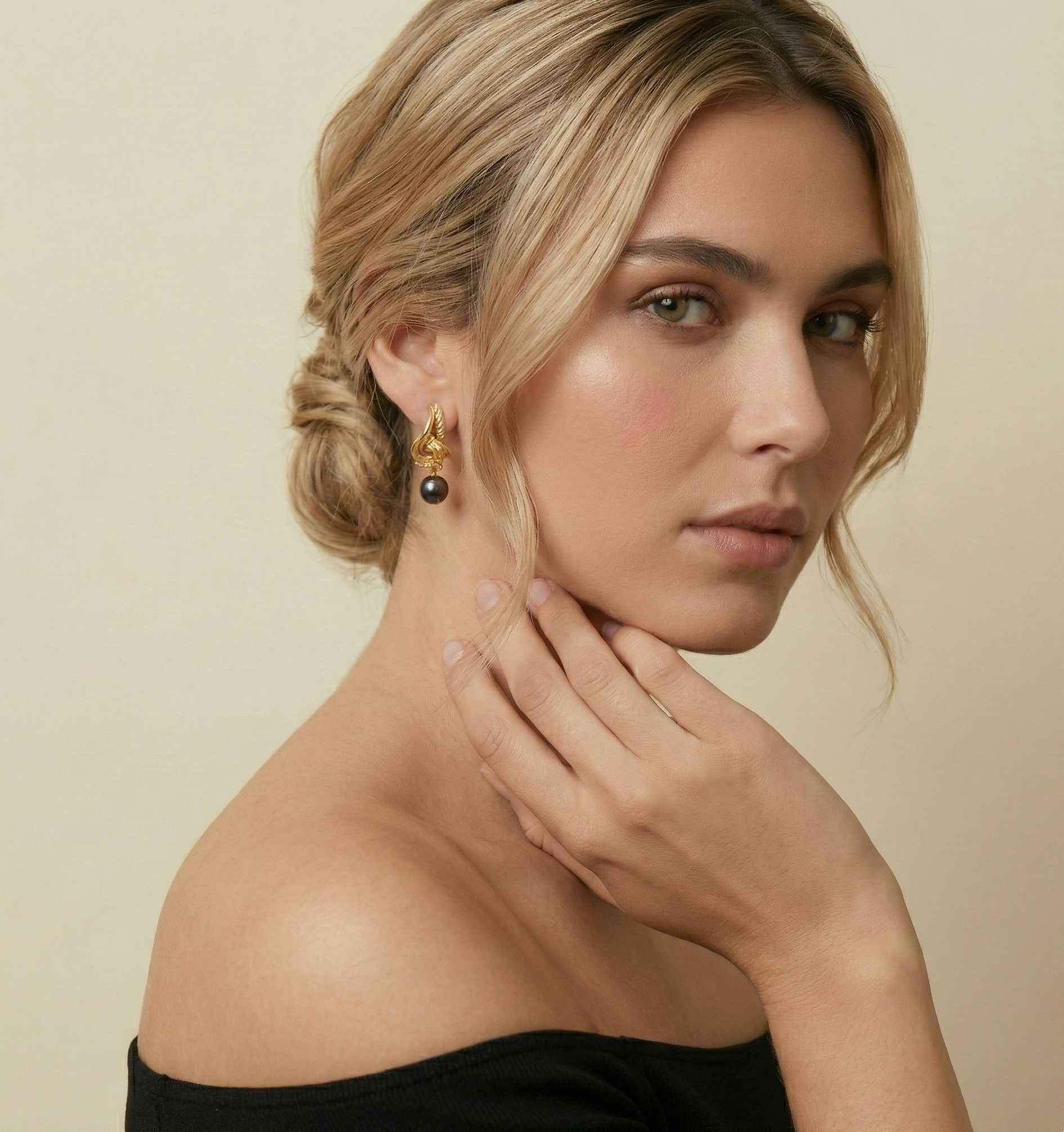 Close-up portrait of a female model wearing elegant gold twisted hoop earrings featuring a lustrous black pearl drop. She has a sophisticated updo hairstyle and wears a black off-shoulder top, highlighting the jewelry against a neutral background