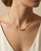 Close-up editorial portrait of a French model wearing a delicate gold chain necklace featuring three small, oval pink pearls with a high silvery luster, styled with a deep-neck ivory silk top against a matte beige background