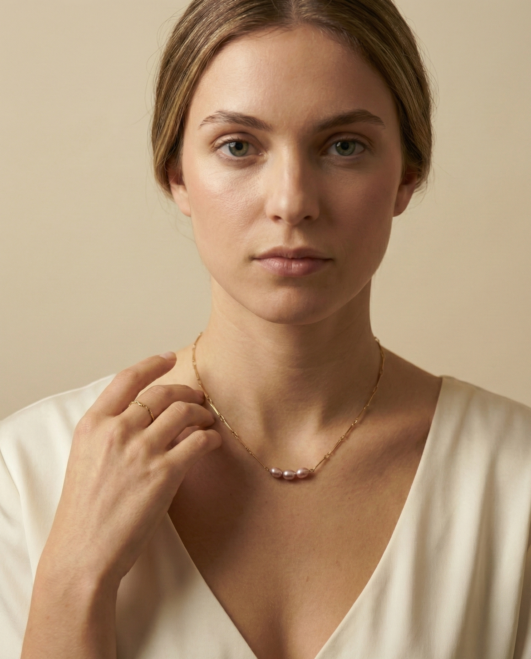 Close-up editorial portrait of a French model wearing a delicate gold chain necklace featuring three small, oval pink pearls with a high silvery luster, styled with a deep-neck ivory silk top against a matte beige background