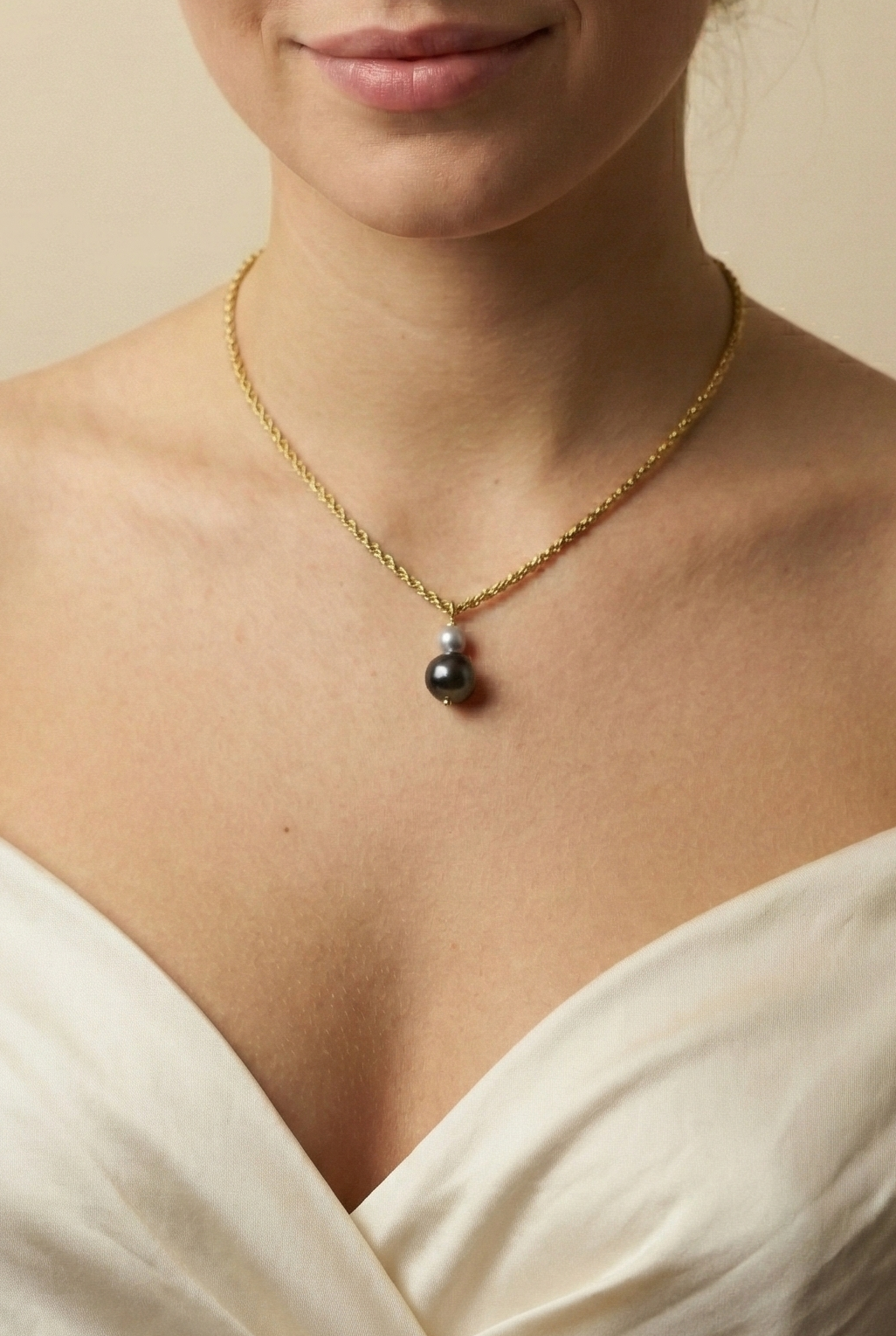 Elegant model with brown-blonde hair wearing a dainty gold rope chain necklace with a black and grey pearl pendant, styled with an off-shoulder silk top