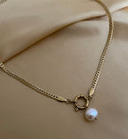 Close-up detail of a flat gold curb chain necklace featuring a lustrous white baroque pearl pendant attached to a round front spring clasp