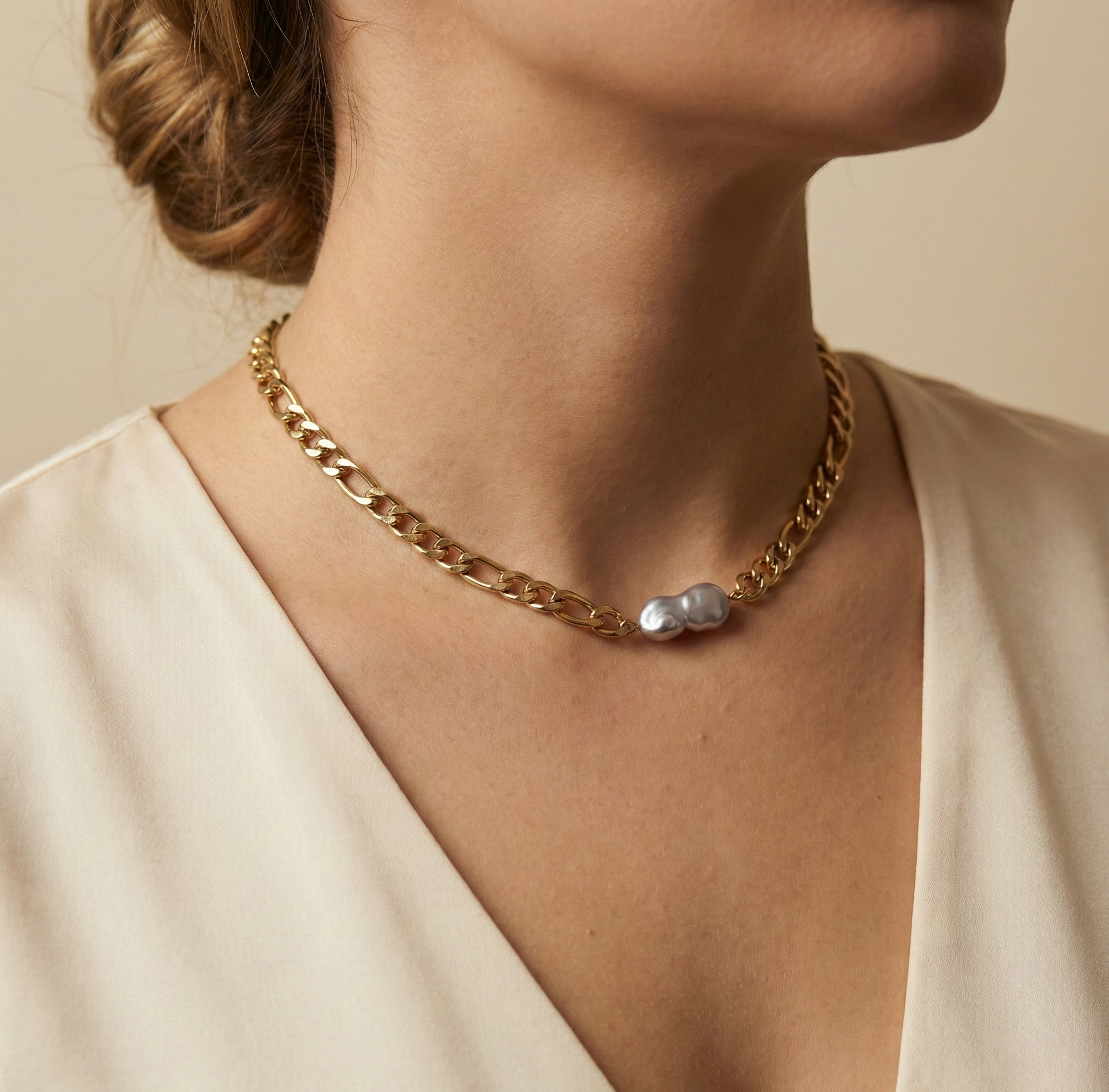 Close-up editorial portrait of a woman wearing a gold Figaro chain necklace featuring a unique grey Keshi baroque pearl with metallic luster. The necklace sits on the collarbone, styled with a deep V-neck cream silk top