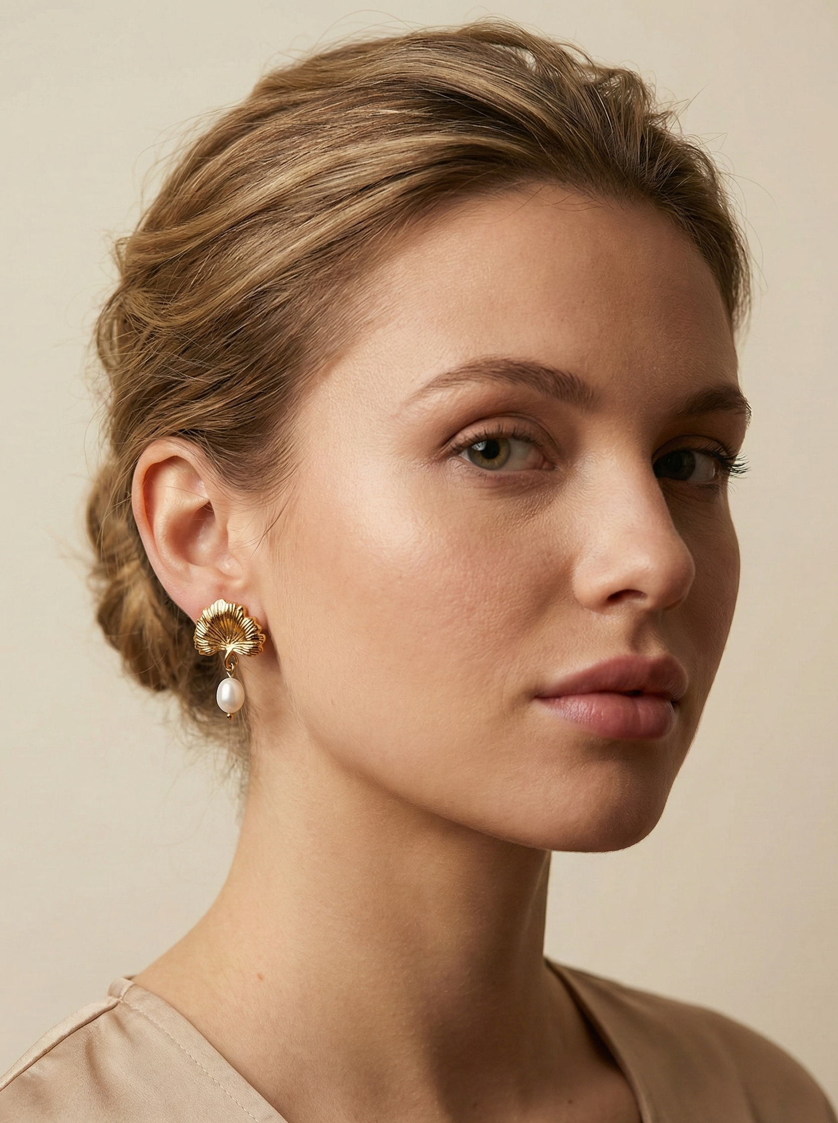 Close-up editorial jewelry photography of a woman wearing gold ginkgo leaf stud earrings with white pearl drops. The model has blonde-brown hair styled in a low bun and wears a beige camisole, capturing a chic French aesthetic. The earrings feature a detailed, textured gold fan shape resembling a ginkgo biloba leaf, from which hangs a lustrous white freshwater pearl.