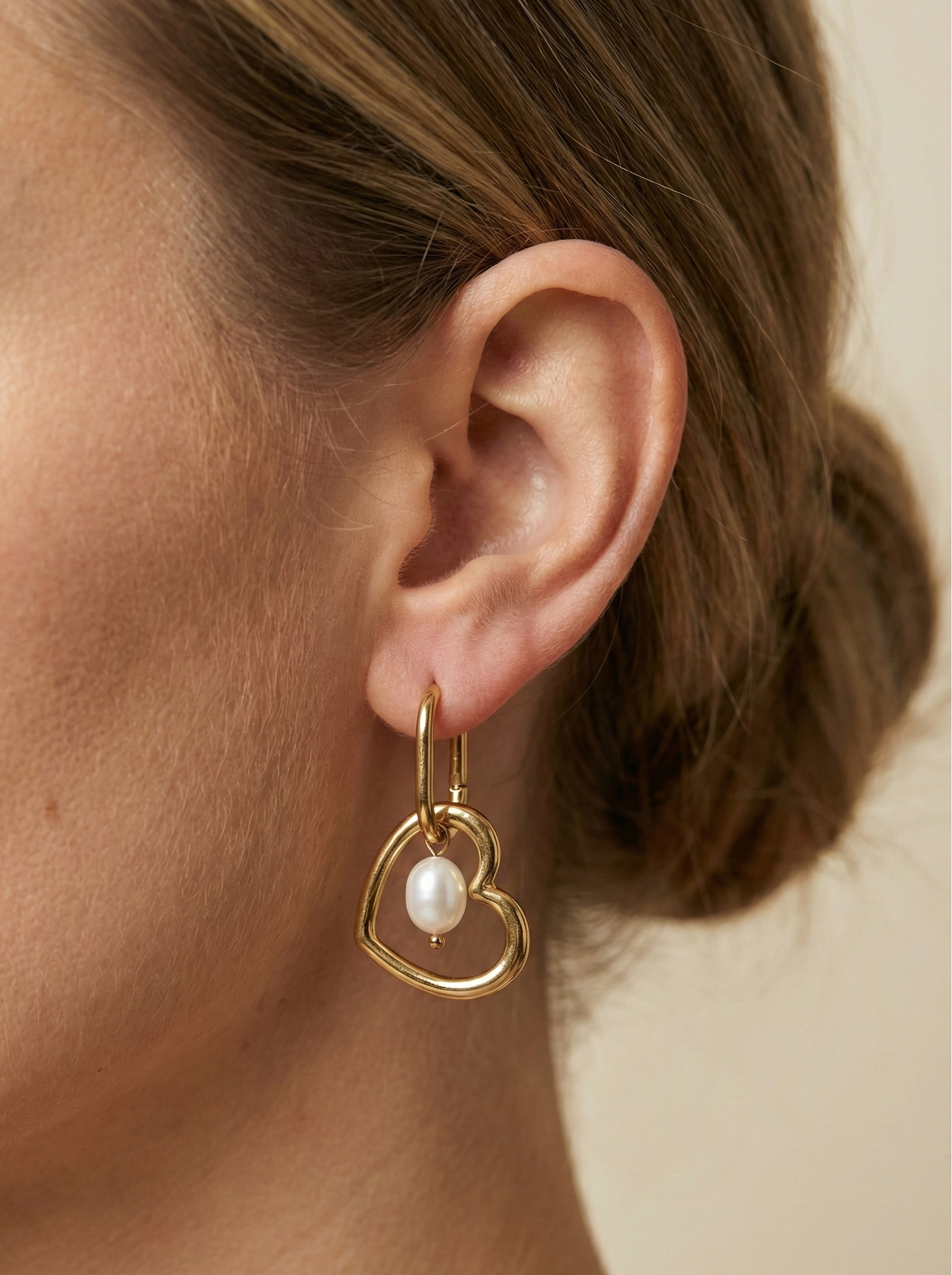Close-up of a model wearing gold heart-shaped hoop earrings with a lustrous white pearl charm. High-quality jewelry photography showcasing the polished gold finish and pearl texture against a plain ivory background