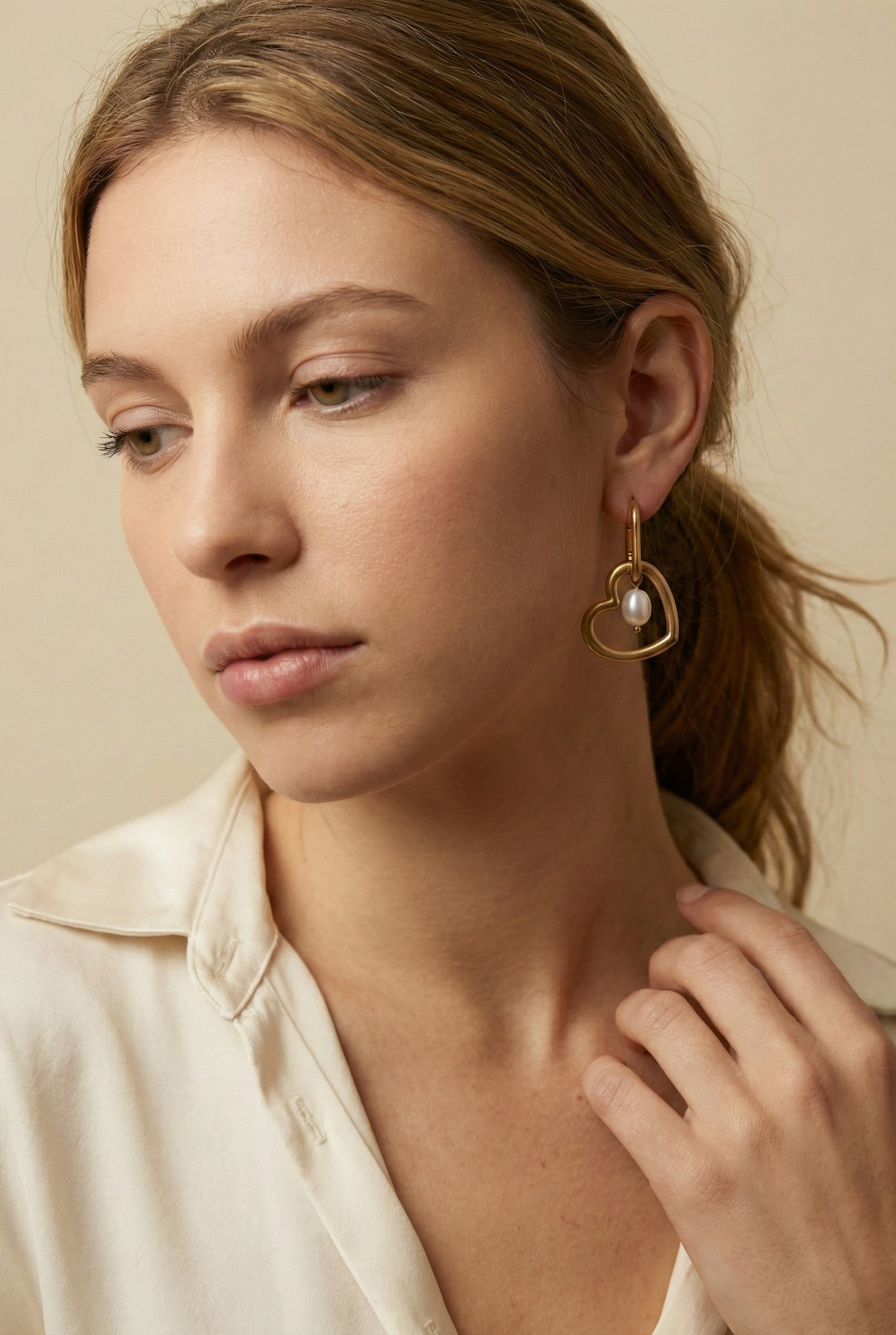 Close-up of a model wearing gold heart-shaped hoop earrings with a lustrous white pearl charm. High-quality jewelry photography showcasing the polished gold finish and pearl texture against a plain ivory background