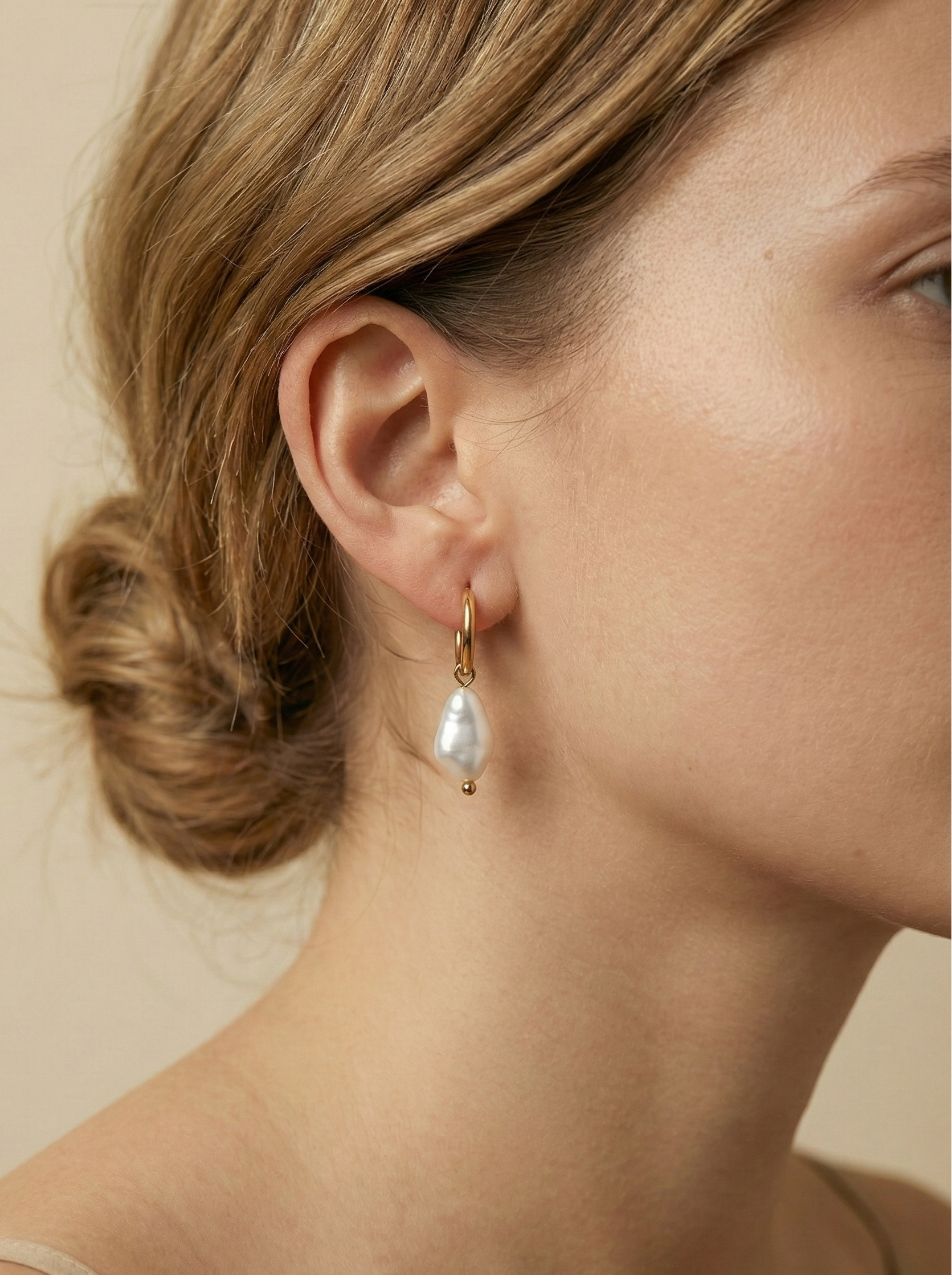 Close-up photography of a dainty gold hoop earring featuring a lustrous white baroque pearl drop worn on a model's ear. High-resolution commercial jewelry shot showcasing organic pearl texture, nacre iridescence, and a simple gold clasp against a matte ivory background.