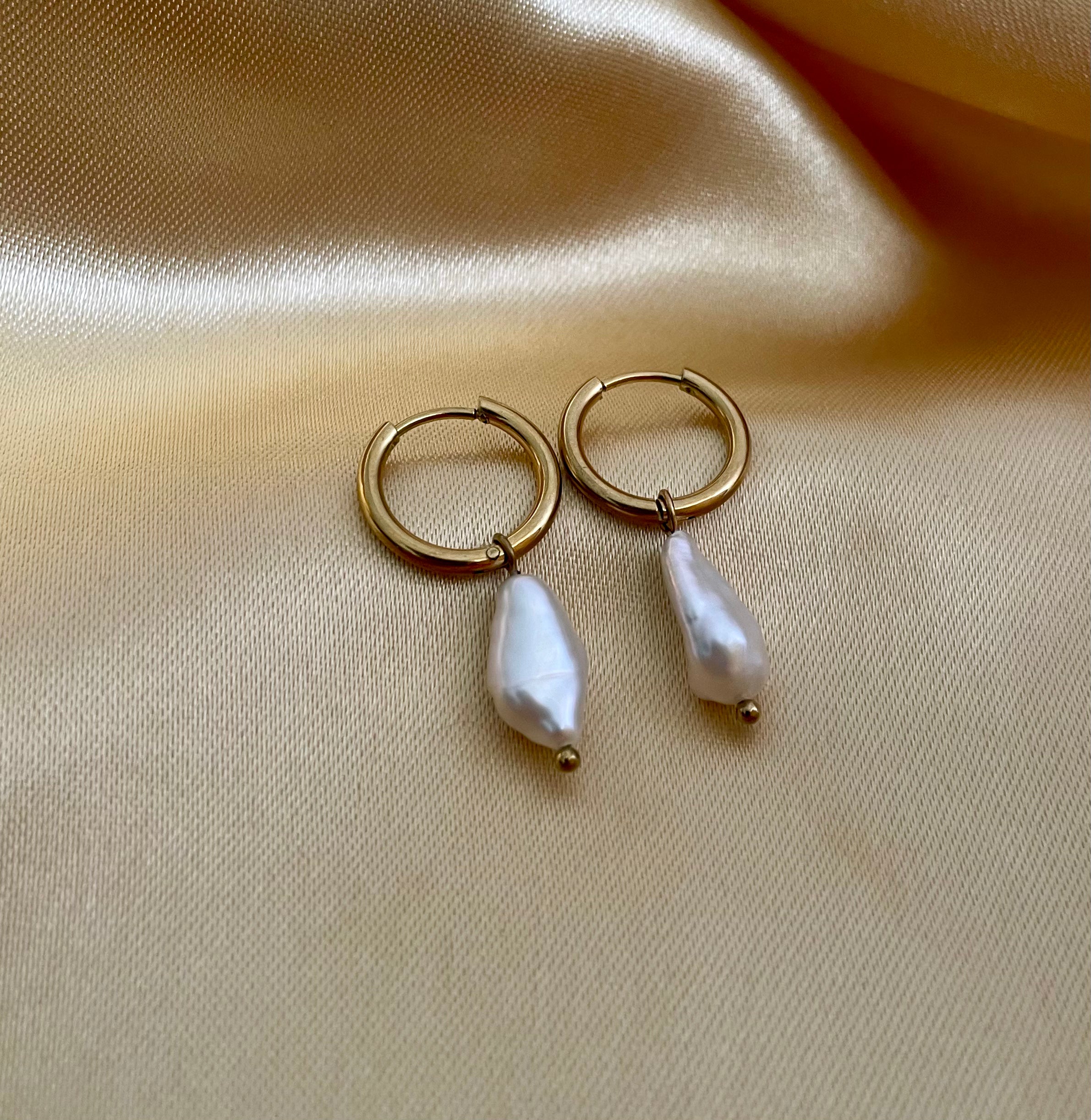 Close-up photography of a dainty gold hoop earring featuring a lustrous white baroque pearl drop worn on a model's ear. High-resolution commercial jewelry shot showcasing organic pearl texture, nacre iridescence, and a simple gold clasp against a matte ivory background.