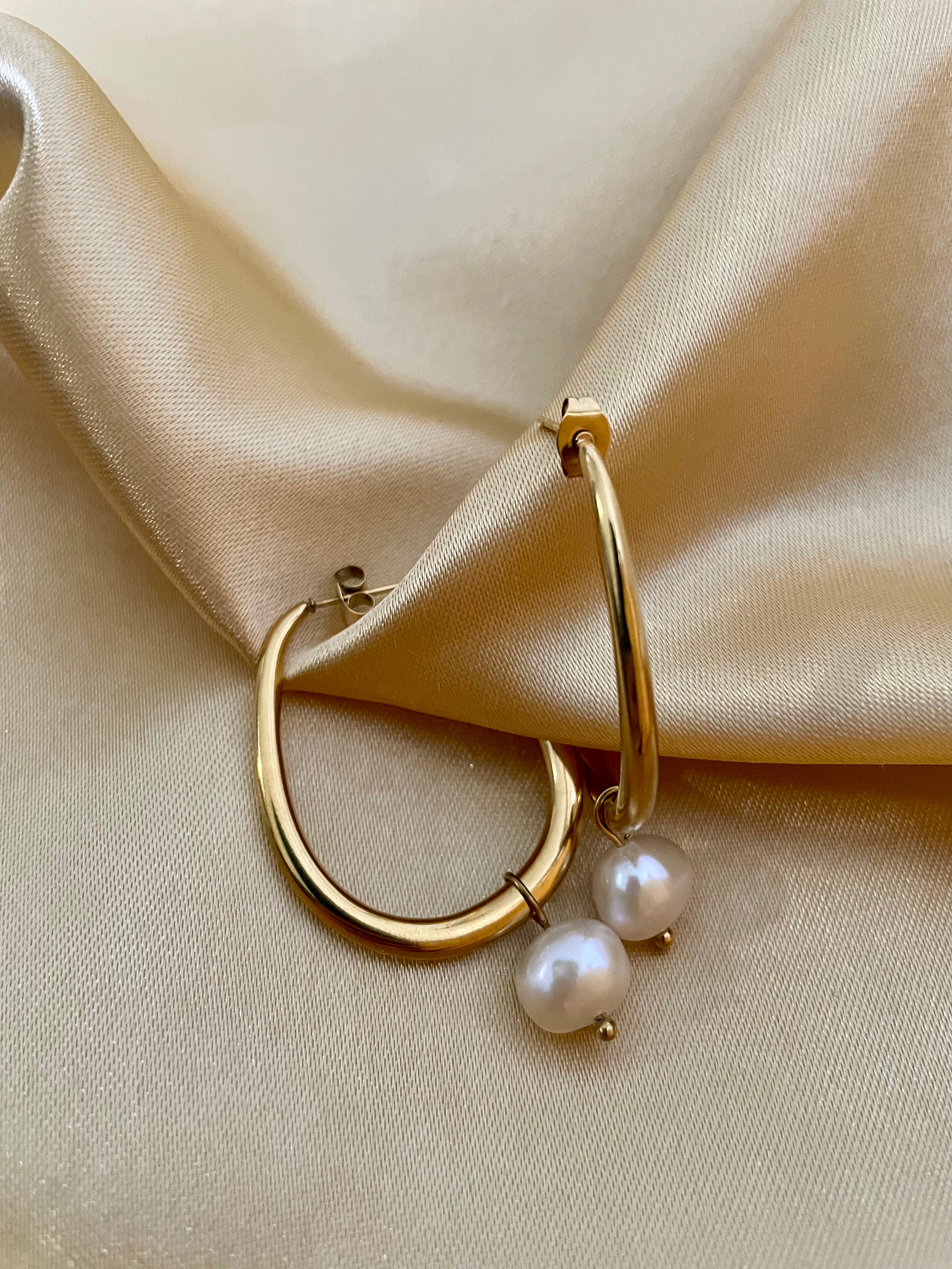 Close-up side profile of a woman's ear wearing a gold hoop earring with a single white pearl drop, highlighting the metallic luster and pearl detail against a soft neutral background.