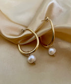 Close-up of a gold hoop earring with a single white pearl drop, highlighting the metallic luster and pearl detail against a soft neutral background