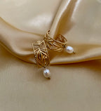 Close-up of a detailed gold openwork hoop earrings featuring a leaf motif and a single dangling white oval pearl. The warm, soft lighting highlights the pearl's luster and the gold texture, set against a neutral background.