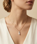 Close-up editorial portrait of a woman wearing a delicate gold chain necklace featuring a stacked pendant with a lustrous white keshi pearl and a small grey baroque pearl, styled with an ivory silk top.