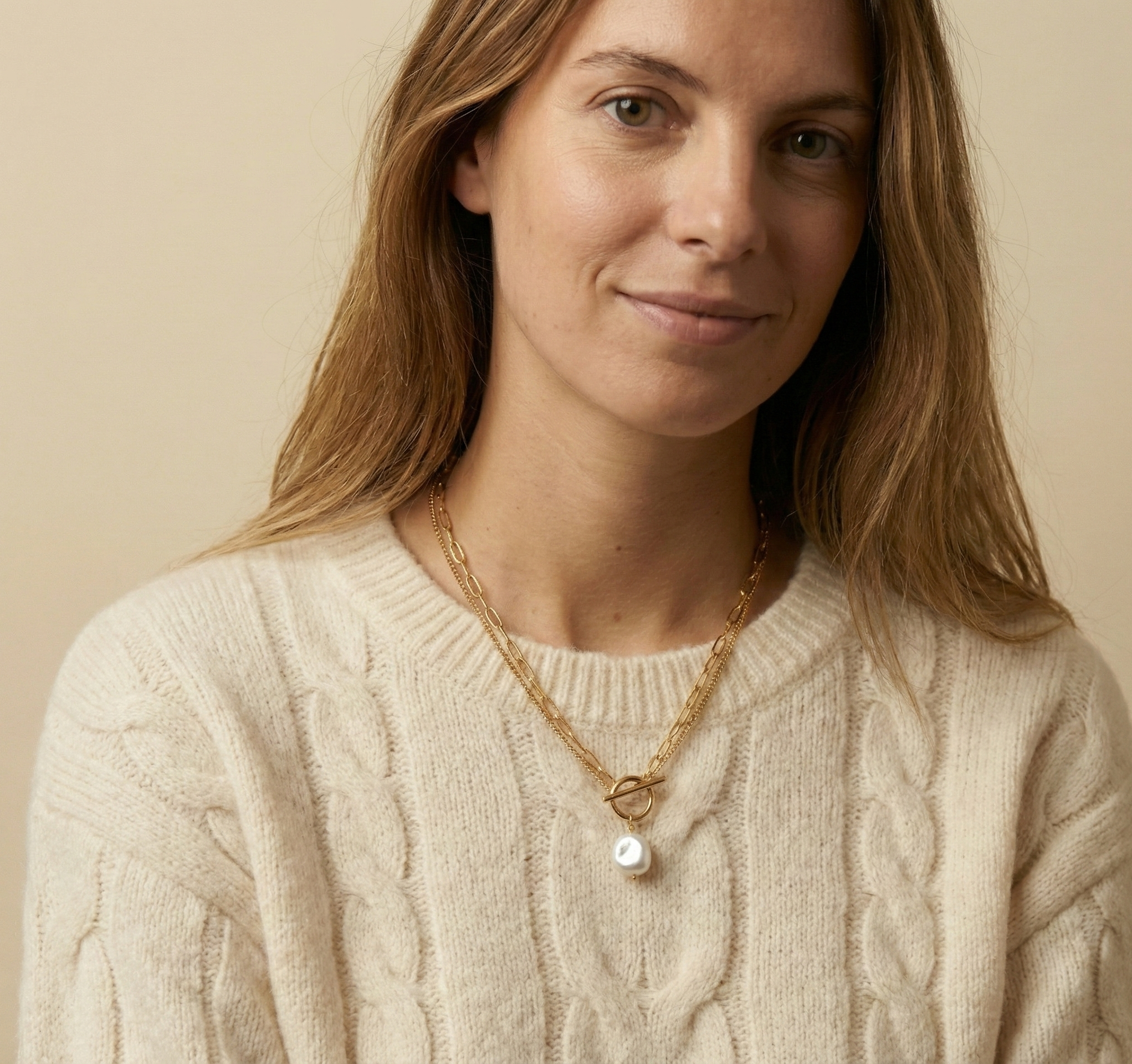 Close-up detail of a double-strand gold chain necklace featuring a lustrous white baroque pearl pendant and a T-bar toggle clasp, worn on the collarbone of a model in a white sweater