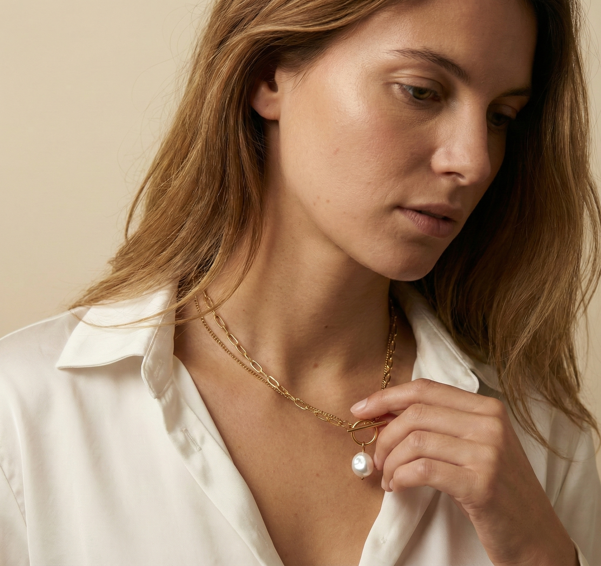 Close-up detail of a double-strand gold chain necklace featuring a lustrous white baroque pearl pendant and a T-bar toggle clasp, worn on the collarbone of a model in an open white silk shirt