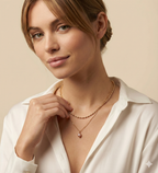 Close-up of a model wearing a layered gold necklace set featuring a choker with small gold beads and a delicate chain with a single dark pink oval pearl pendant, styled with a deep V-neck white silk shirt