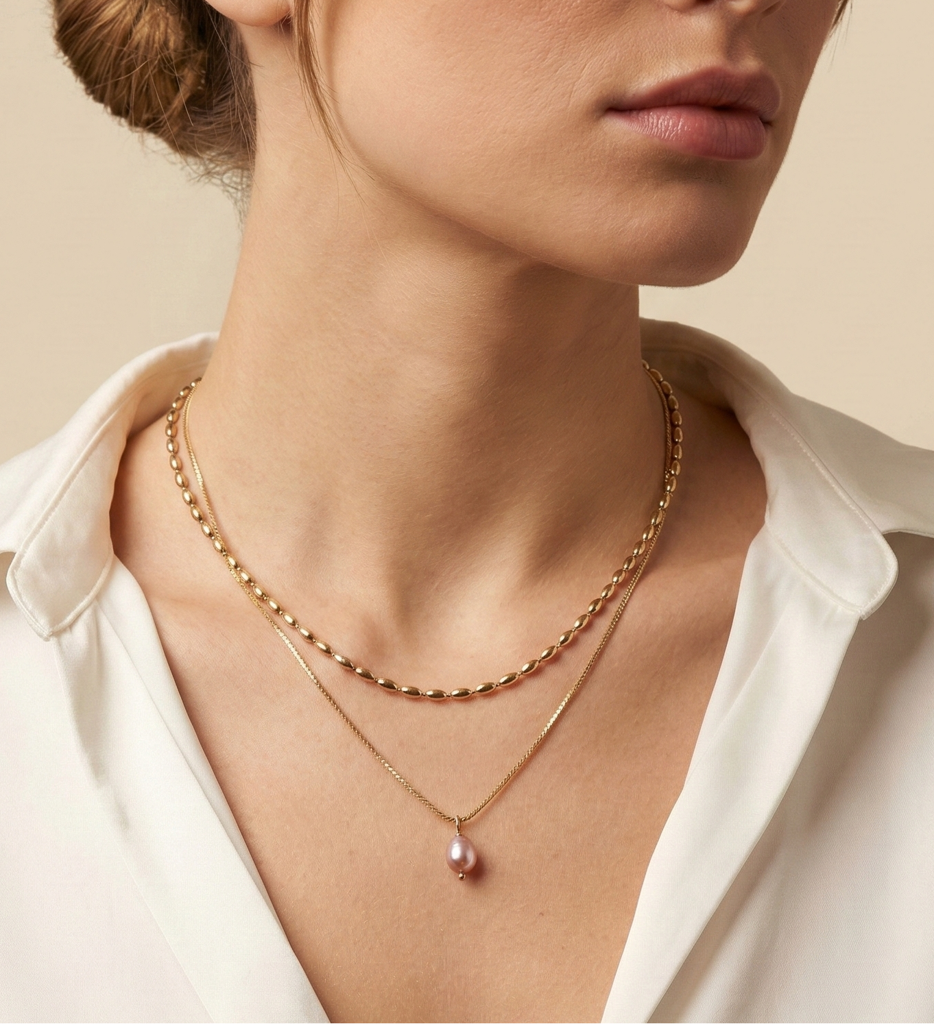 Close-up of a model wearing a layered gold necklace set featuring a choker with small gold beads and a delicate chain with a single dark pink oval pearl pendant, styled with a deep V-neck white silk shirt