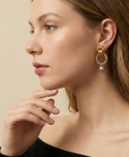 A model with blonde hair wearing elegant 18k gold plated link earrings with a grey pearl drop, styled with a minimalist black top for a luxury editorial look