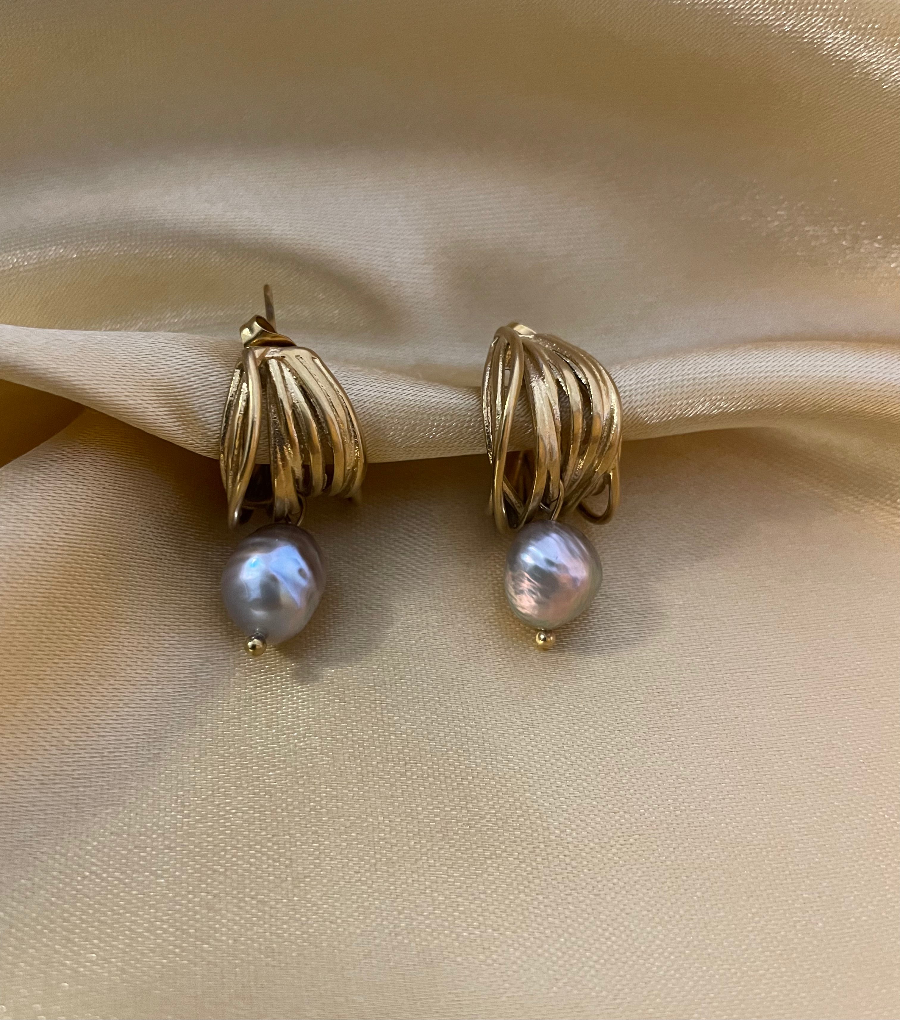 Close-up of 18k gold multi-hoop earrings featuring a lustrous grey pearl drop. The jewelry highlights a textured gold finish and organic pearl shape, set against a plain ivory background