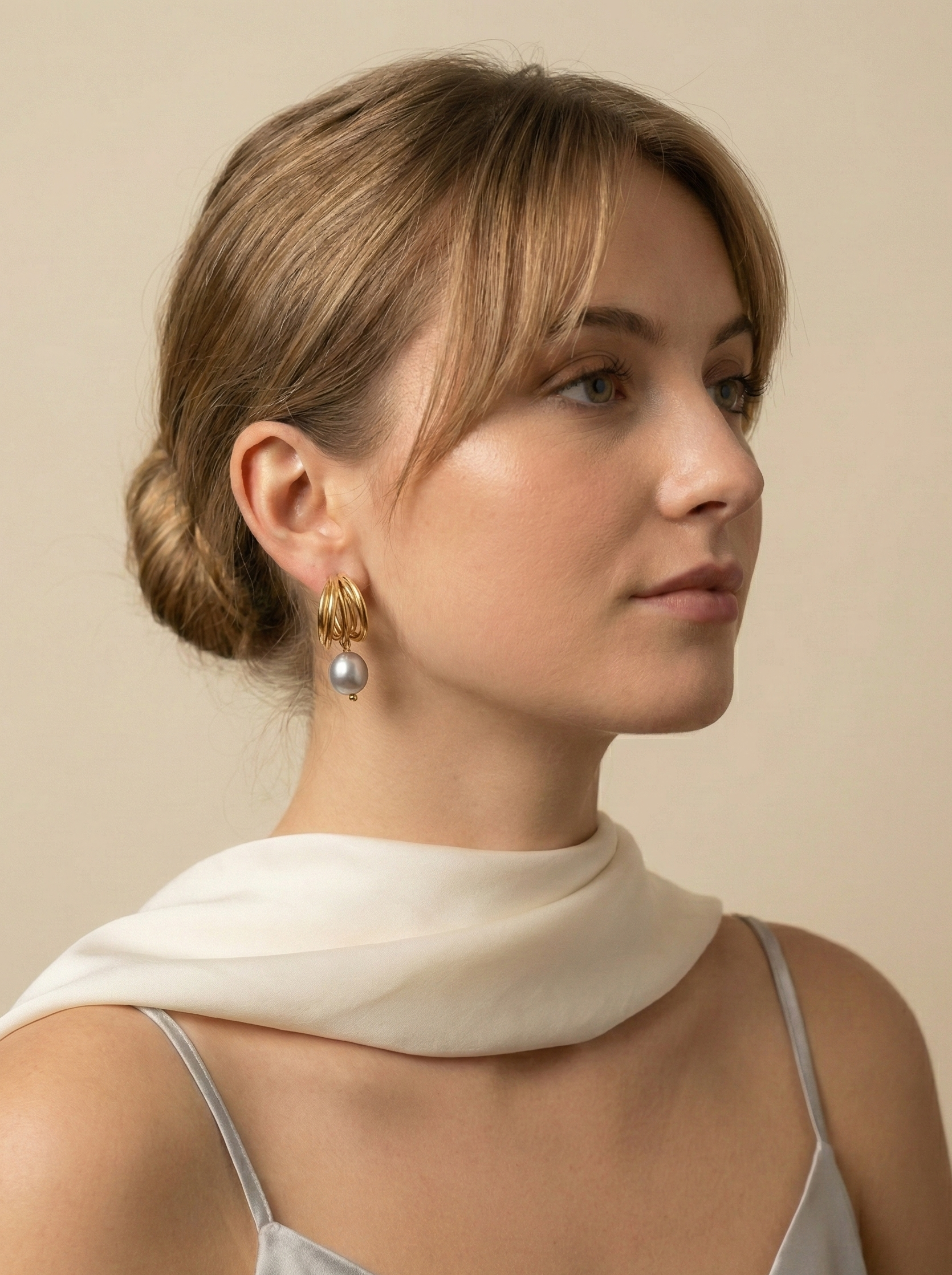 Close-up of 18k gold multi-hoop earrings featuring a lustrous grey pearl drop, worn by a model with a low bun hairstyle. The jewelry highlights a textured gold finish and organic pearl shape, set against a plain ivory background.