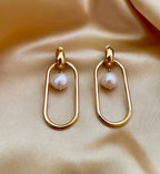 Close-up view of gold elongated oval hoop earrings featuring a suspended white baroque pearl, shown worn on the ear to verify size and scale