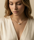 Close-up of a model wearing a gold paperclip chain necklace featuring a single white baroque pearl pendant, styled with a cream silk deep V-neck top