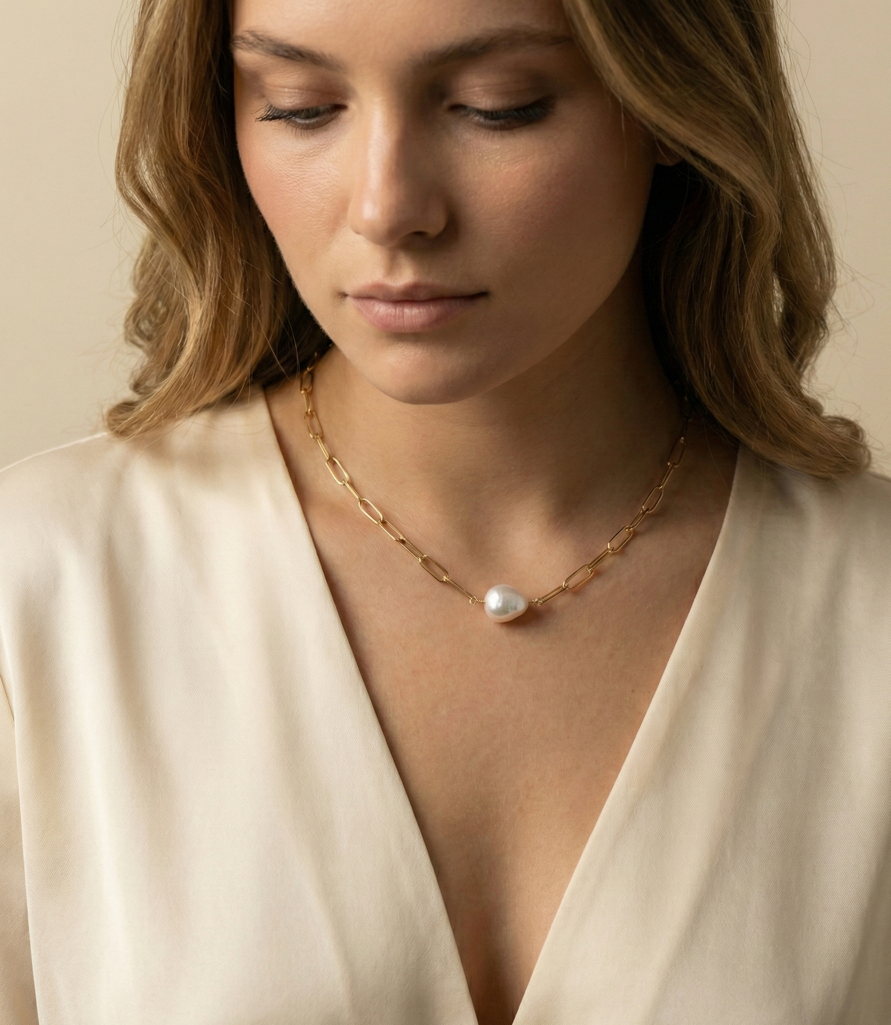 Close-up of a model wearing a gold paperclip chain necklace featuring a single white baroque pearl pendant, styled with a cream silk deep V-neck top