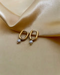 Close-up of gold rectangular huggie hoop earrings featuring a dainty grey pearl drop charm, worn on the ear.