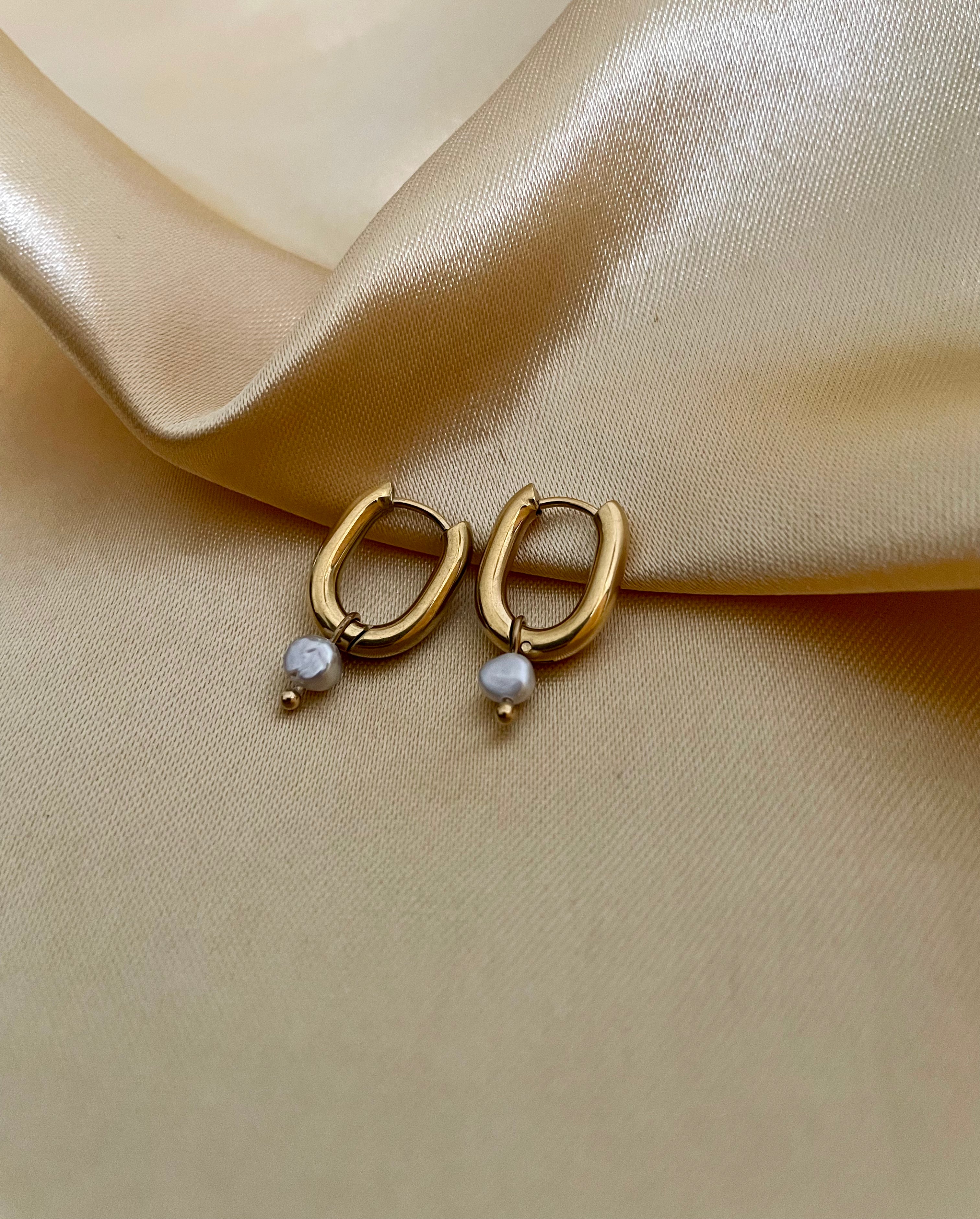 Close-up of gold rectangular huggie hoop earrings featuring a dainty grey pearl drop charm, worn on the ear.