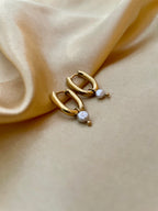 Close-up of gold rectangular huggie hoop earrings featuring a dainty grey pearl drop charm