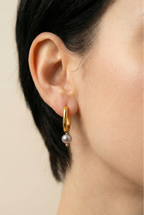 Close-up of gold rectangular huggie hoop earrings featuring a dainty grey pearl drop charm, worn on the ear