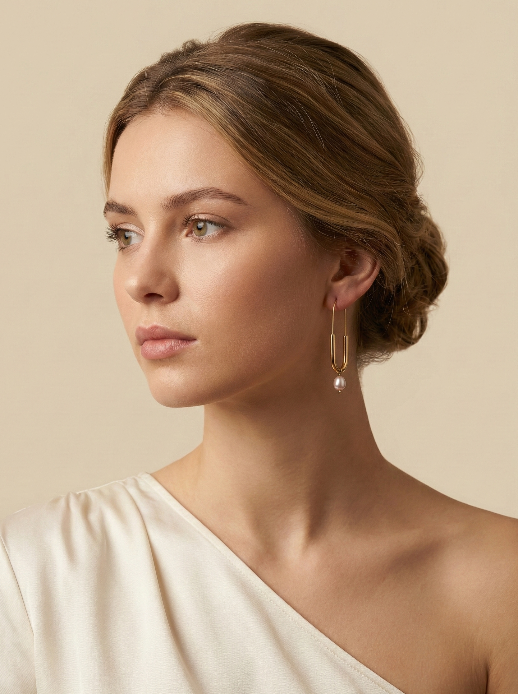 Close-up of a model wearing modern gold safety pin drop earrings featuring a freshwater lustrous pink oval pearl charm. Minimalist gold wire earrings with a U-shape design and natural pearl detail