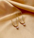 Close-up of a model wearing modern gold safety pin drop earrings featuring a lustrous pink oval pearl charm. Minimalist gold wire earrings with a U-shape design and natural pearl detail