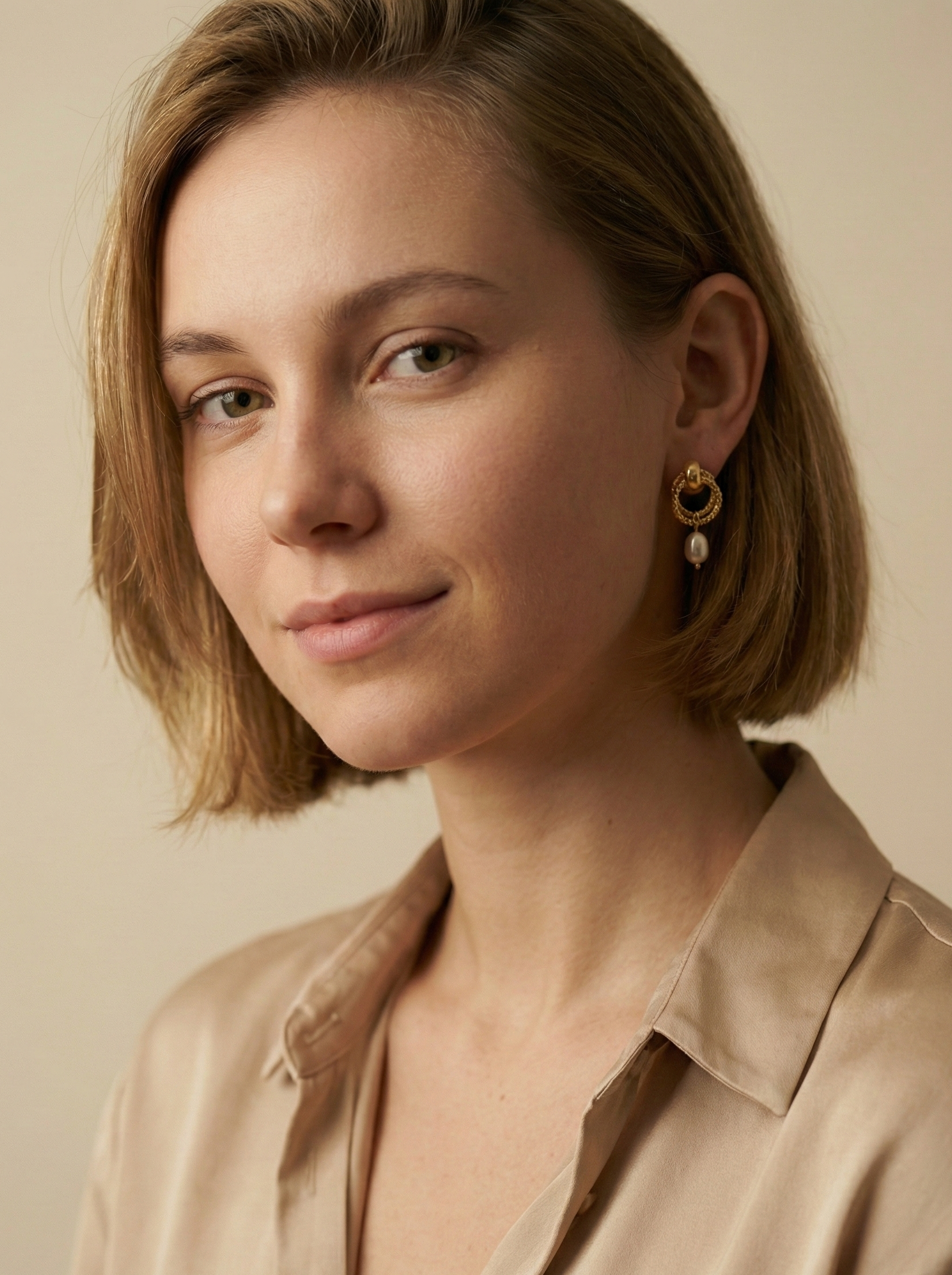 Close-up detail of textured gold hoop earrings featuring a dainty oval pearl drop. The earrings are worn on the ear to show scale, highlighting the metallic luster and elegant Parisian chic design against a neutral background.