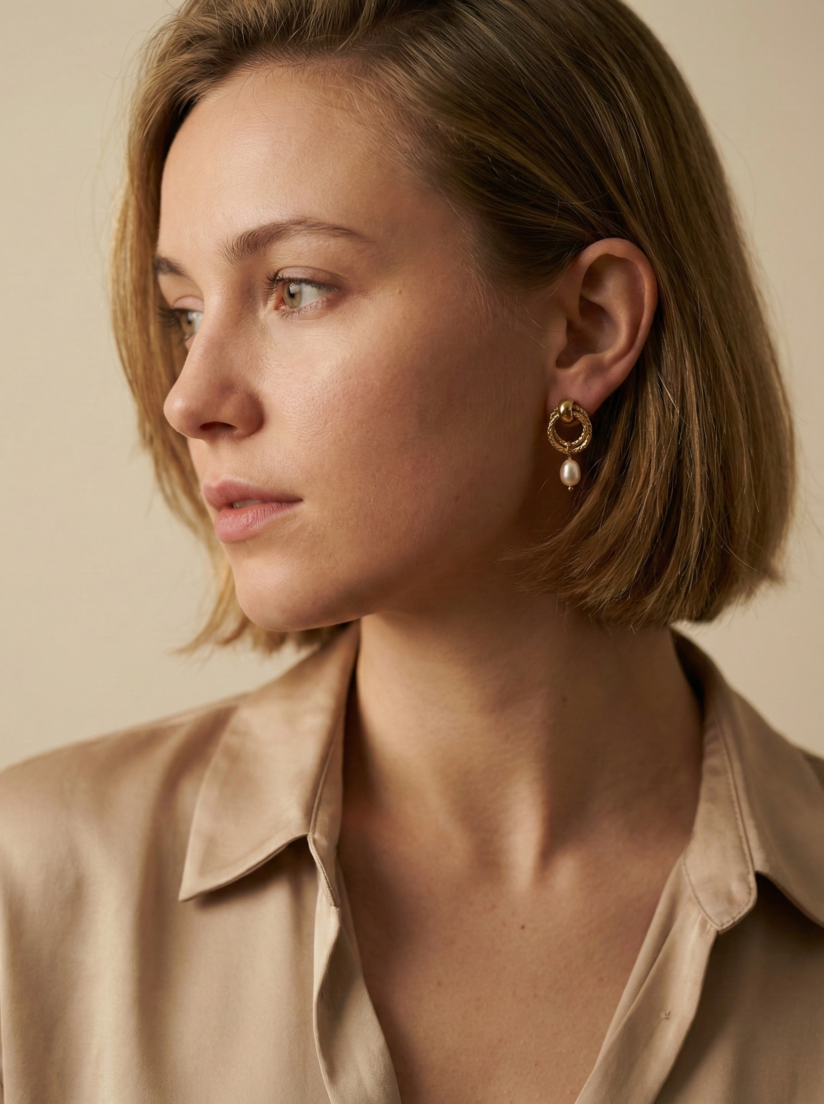 Close-up detail of textured gold hoop earrings featuring a dainty oval pearl drop. The earrings are worn on the ear to show scale, highlighting the metallic luster and elegant Parisian chic design against a neutral background.