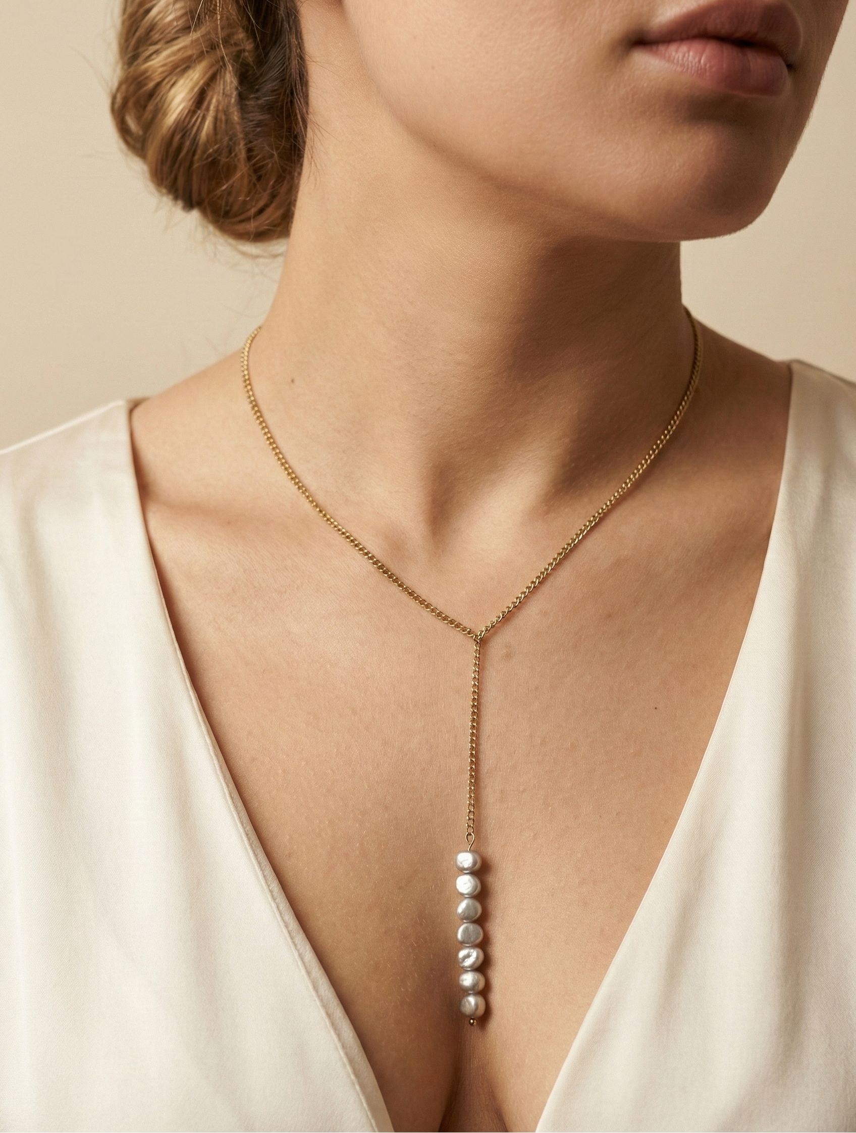 Close-up of a model wearing a delicate gold Y-necklace featuring a vertical drop of seven lustrous grey baroque pearls, styled with a deep V-neck ivory silk top