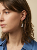 Close-up profile of a french model wearing luxury grey baroque pearl drop earrings with a sculptural gold double-oval link stud. The image highlights the iridescent nacre and organic shape of the pearl, styled with a blue knit top against a soft matte ivory background, representing high-end commercial jewelry photography