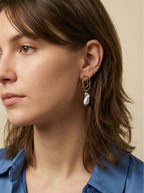 Close-up profile of a french model wearing luxury grey baroque pearl drop earrings with a sculptural gold double-oval link stud. The image highlights the iridescent nacre and organic shape of the pearl, styled with a blue knit top against a soft matte ivory background, representing high-end commercial jewelry photography